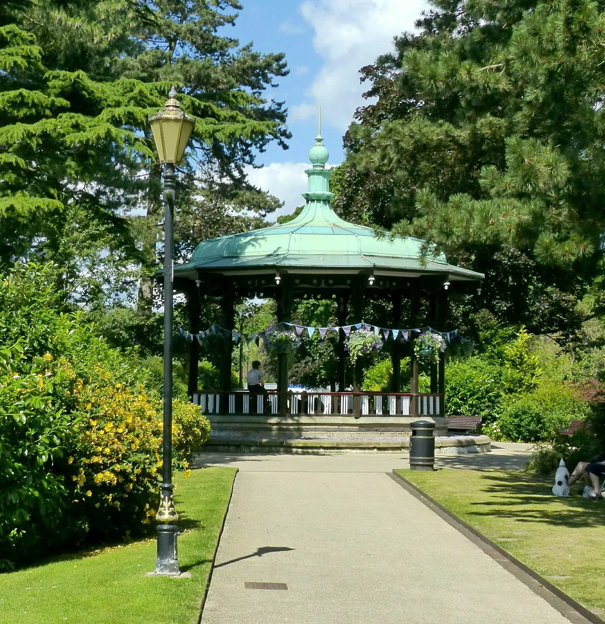 The green copper-roofed bandstand in Belper’s River Gardens on a summer day, hung with bunting and flower baskets, framed by tall trees and a lamp post.